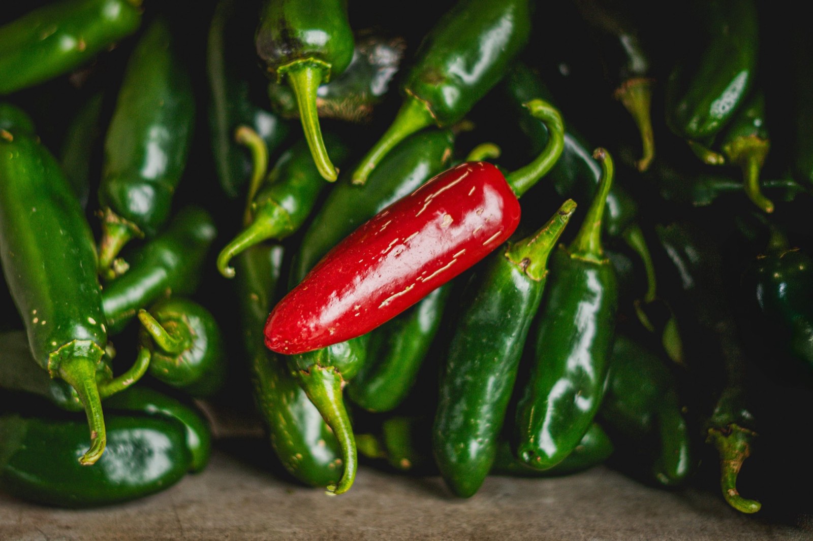 A pile of glossy green jalapeños with a single red jalapeño resting on top.